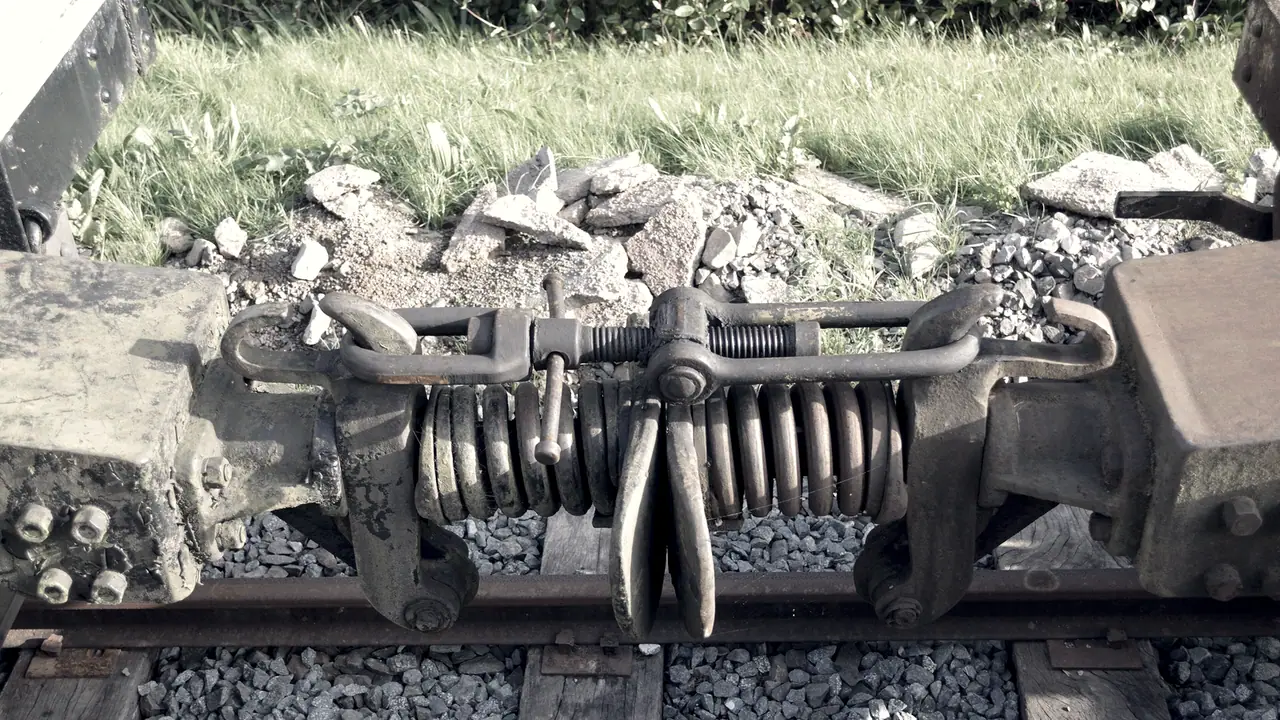 Attelage entre deux wagons, vu au-dessus de la voie : un tendeur à vis relie les deux tampons, au-dessus d'un gros ressort et de deux crochets pendants, sur le ballast.