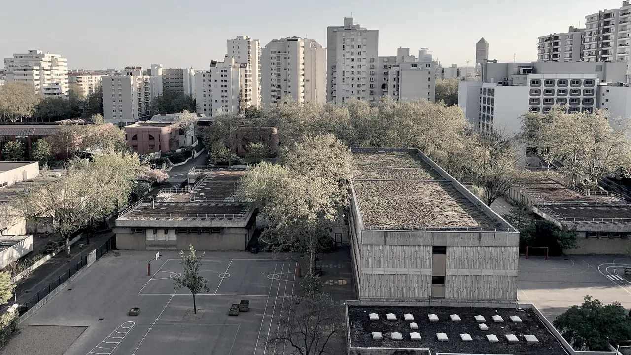 Cour d'école bitumée et bâtiments bas vus d'en haut, avec des tours d'habitation et des arbres à l'arrière-plan.