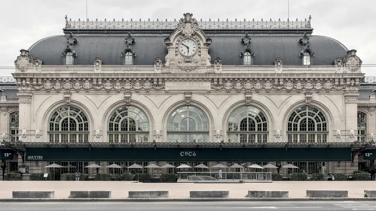 Façade monumentale d'une ancienne gare, avec horloge centrale, verrières cintrées et terrasses sous les auvents.