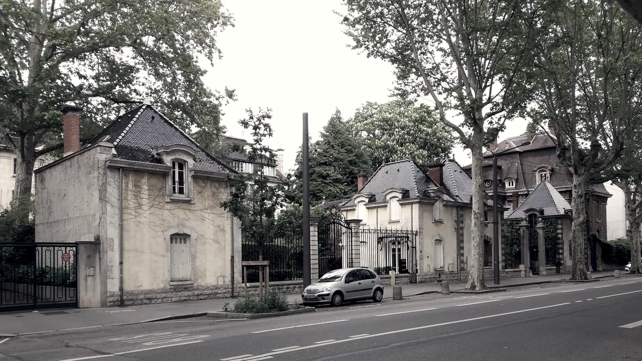 Deux petites maisons anciennes à toit pentu le long d'un boulevard planté de platanes, entre des grilles de fer forgé, avec une villa de brique au fond.