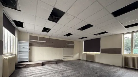 Grande pièce vide avant travaux : dalles de faux plafond déposées, parquet à bâtons rompus usé et radiateurs en fonte sous les fenêtres.