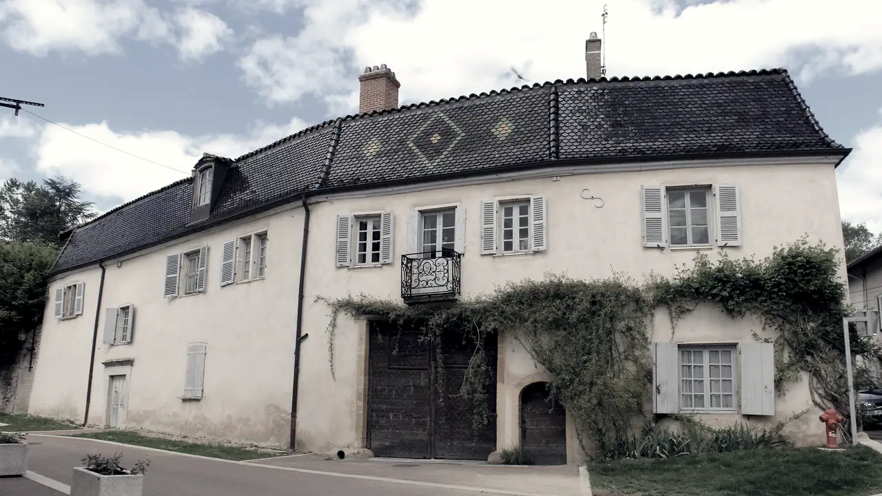 L’ancien relais de poste de Guéreins, dans l’Ain : une longue maison à volets sous un haut toit de tuiles à losanges, avec sa porte cochère en bois.