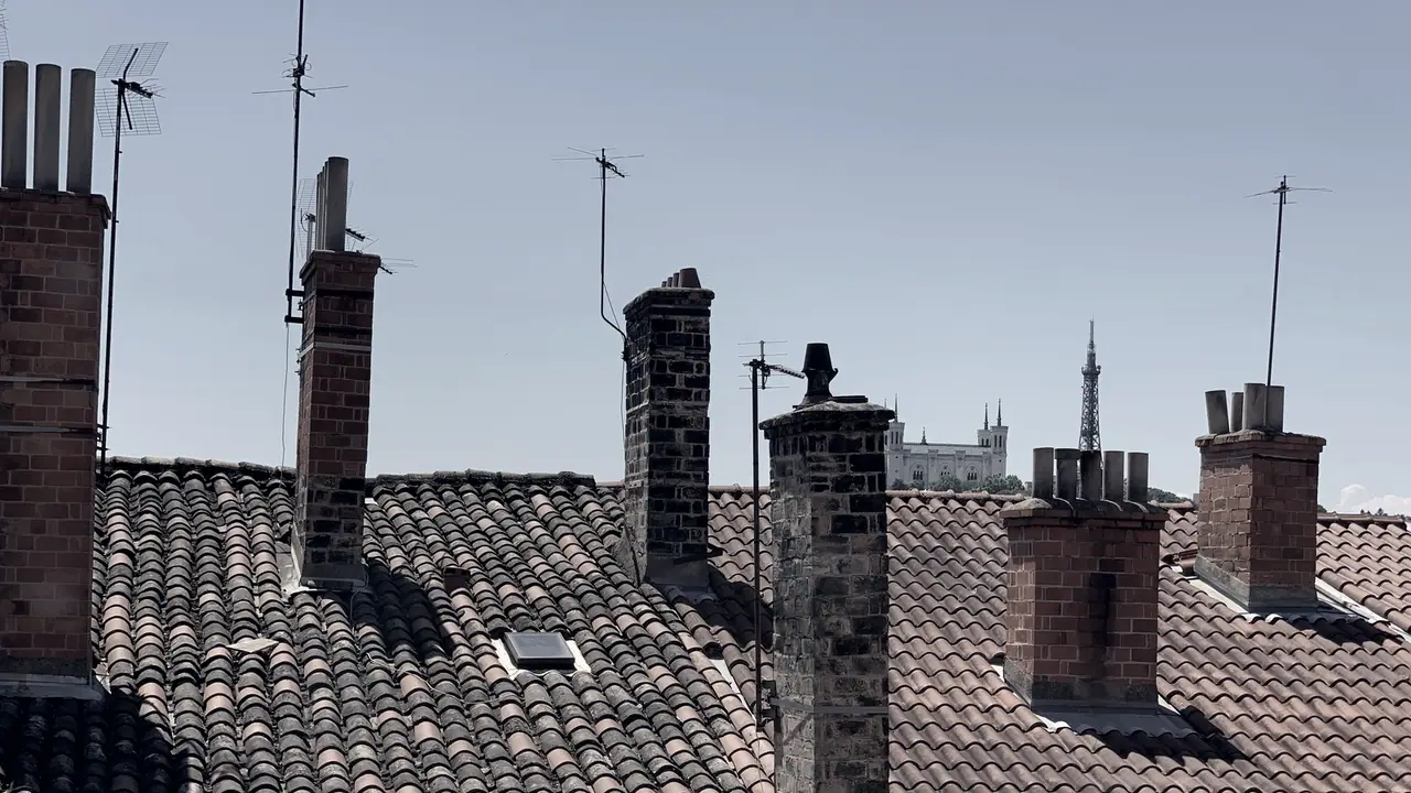 Toits de tuiles rondes et cheminées de brique au premier plan, avec des antennes et une basilique posée sur la colline au loin.