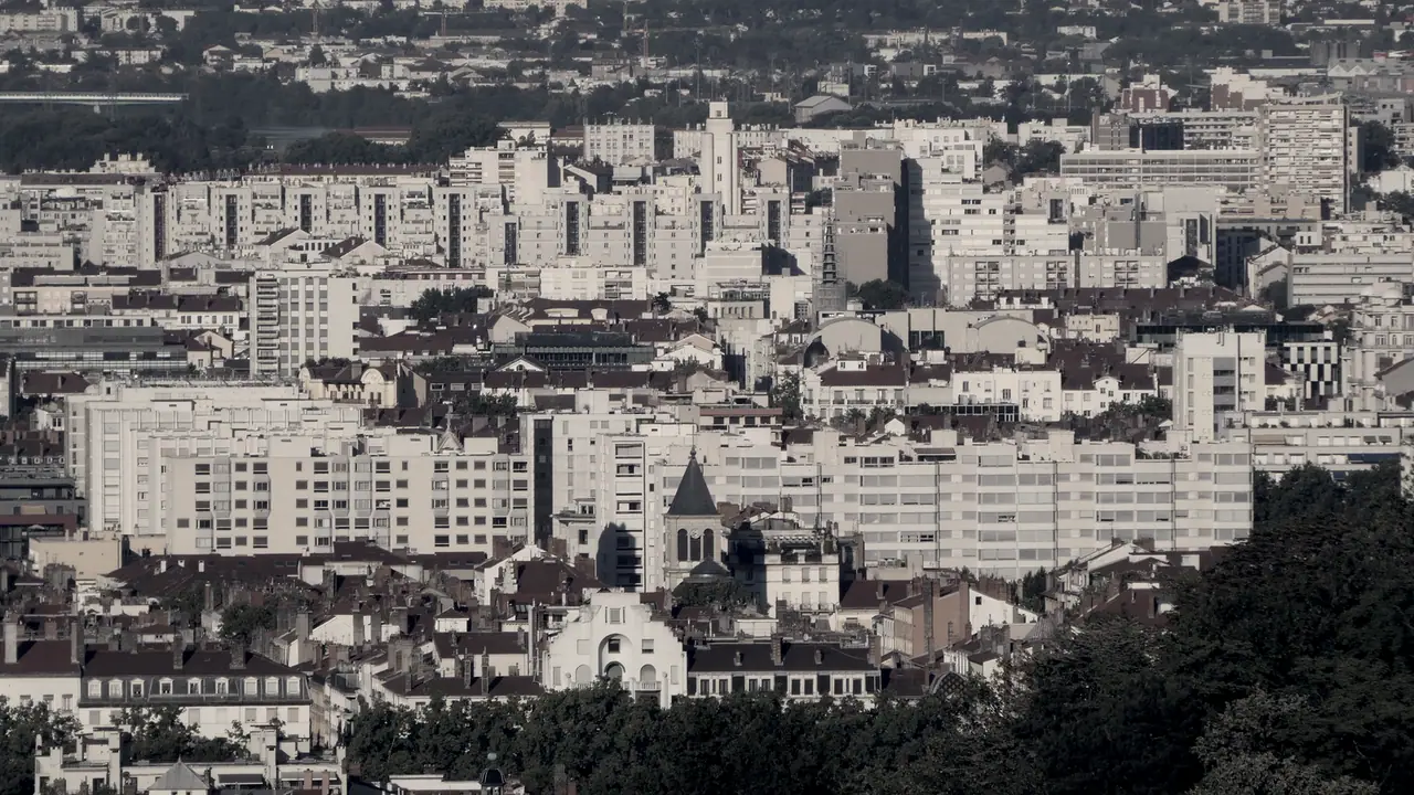 Vue lointaine sur un quartier dense d’immeubles d’habitation de toutes hauteurs, qui s’étend jusqu’au fond du cadre, derrière un clocher.