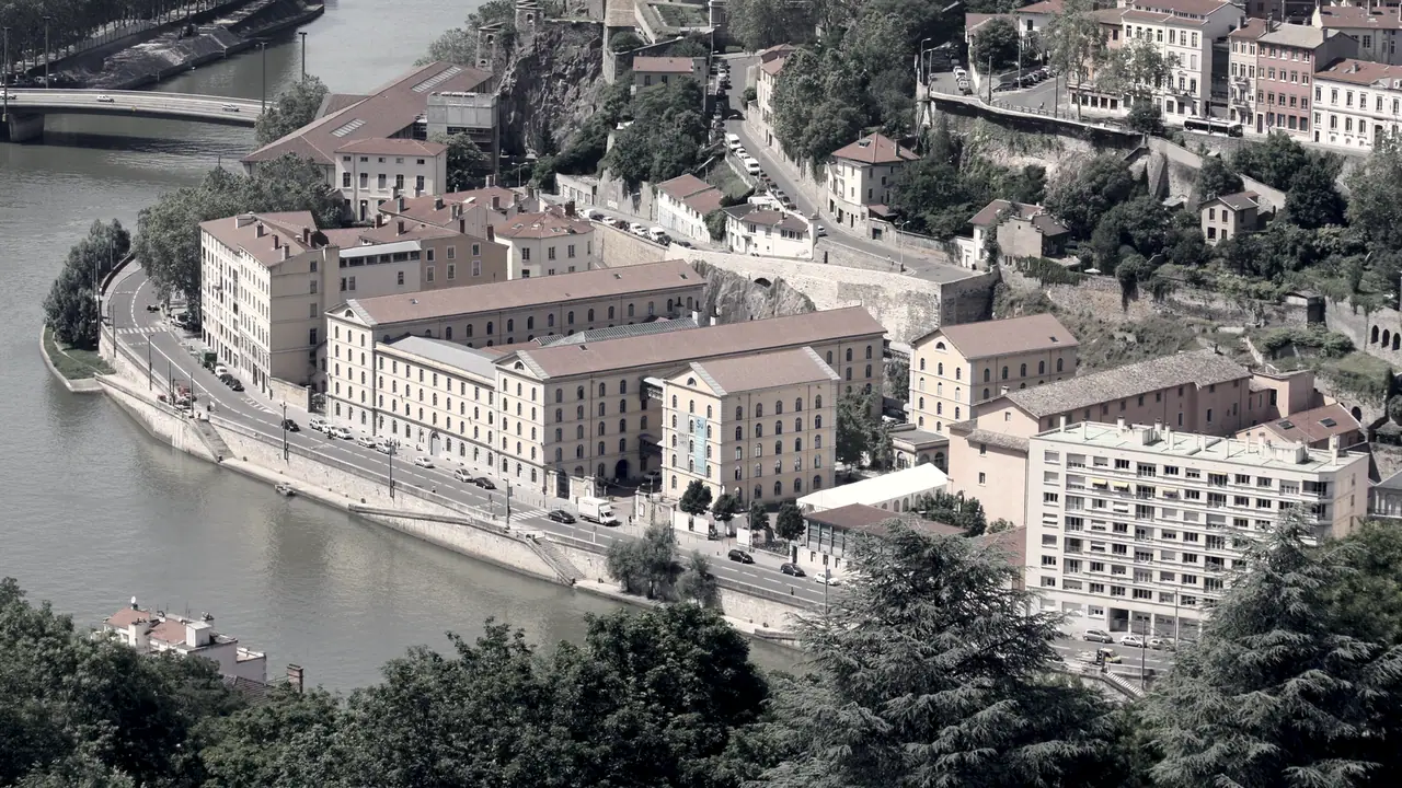 Vue plongeante depuis les toits de Fourvière sur un méandre de la Saône, avec un long bâtiment ancien au bord du quai et des maisons étagées sur la colline.