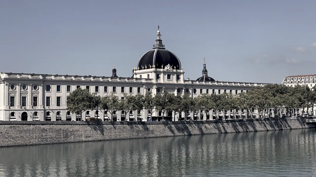 Le Grand Hôtel-Dieu de Lyon vu depuis un pont, sa longue façade à dôme alignée derrière une rangée d’arbres au-dessus du quai du Rhône.