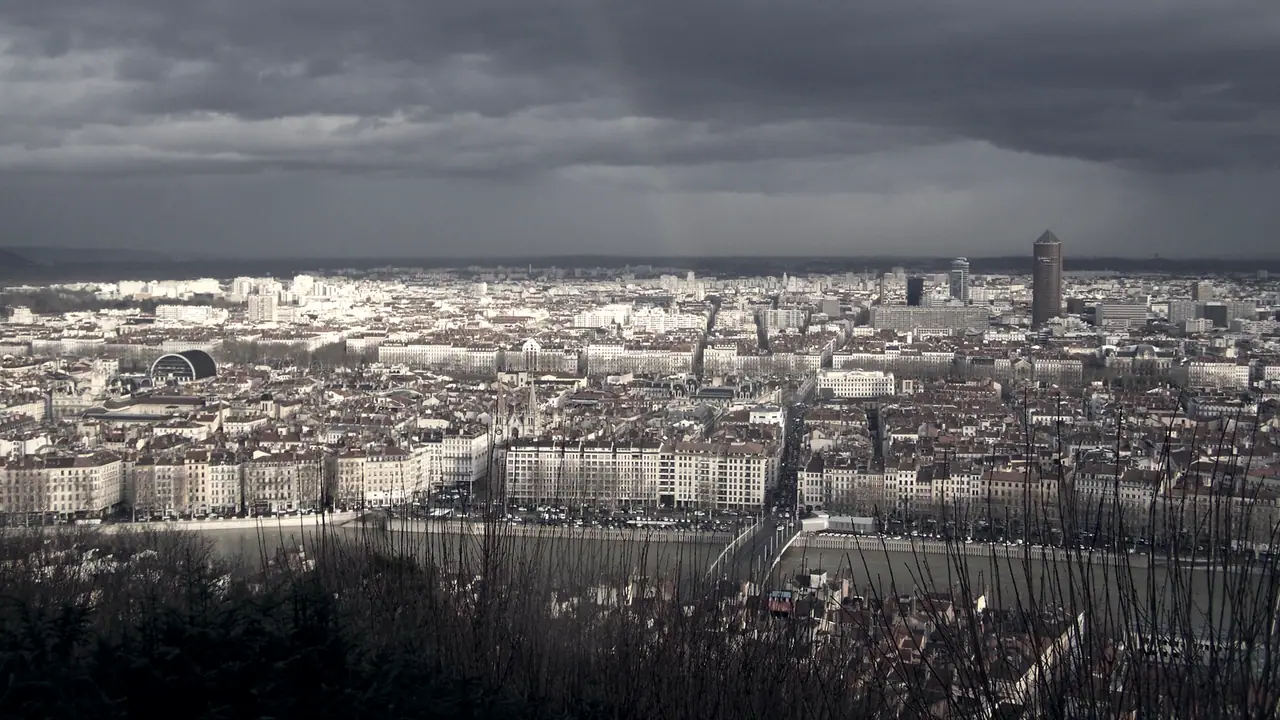 Panorama de Lyon depuis Fourvière sous un ciel d’averse, avec la Saône au premier plan, les toits de la Presqu’île et une haute tour au loin.