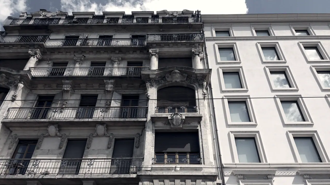 Haut de deux façades mitoyennes le long d’un quai : balcons de fer forgé et consoles sculptées d’un côté, fenêtres régulières et nues de l’autre.