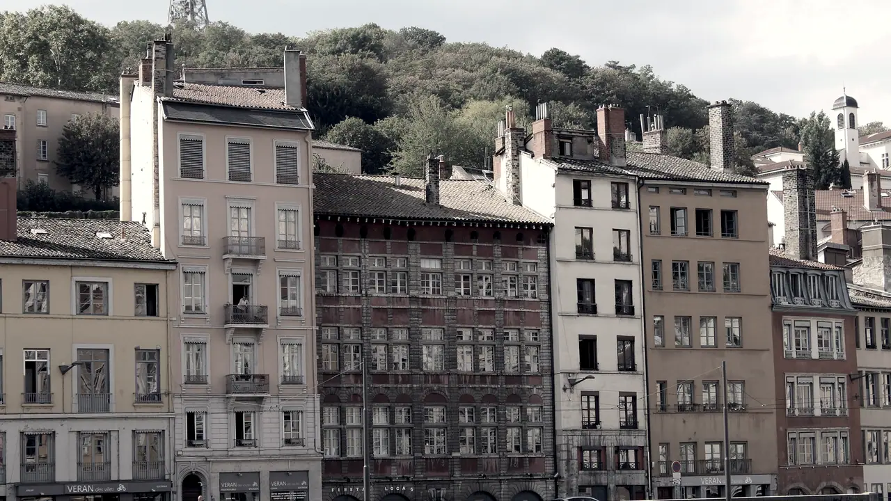 Rangée d’immeubles anciens aux façades étroites et hautes le long d’un quai, au pied d’une colline boisée d’où dépasse un clocheton.