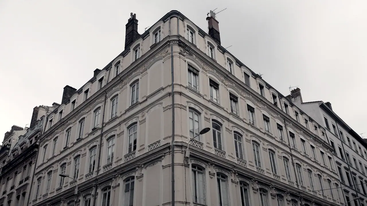 Angle d'un grand immeuble ancien saisi en contre-plongée, avec des rangées de fenêtres à moulures et des cheminées dressées sur le toit.