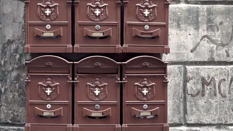 Six boîtes aux lettres en métal ouvragé alignées sur deux rangées contre un mur de pierre décrépi, chacune avec sa serrure et sa fente.