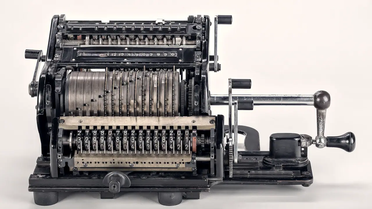 Machine à calculer mécanique ancienne, avec ses rangées de chiffres, ses tambours et sa manivelle, posée sur un fond uni.