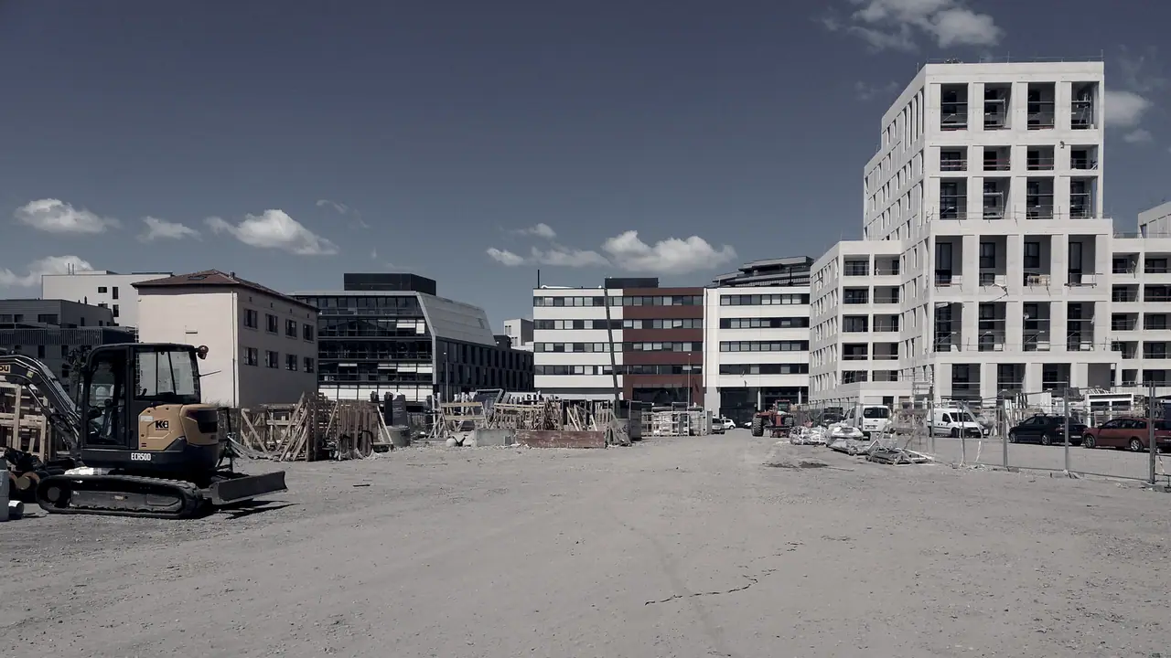Mini-pelle posée sur un terrain de chantier dégagé, devant des immeubles neufs en construction et des palissades.